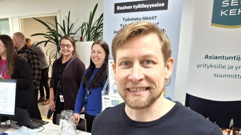 Ilkka Koppelomäki hymyilee. Taustalla TE-asiantuntijoita messuilla.